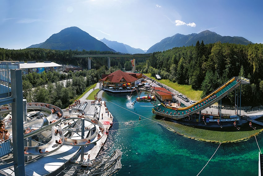 Panoramablick auf das Erlebnisbad Area 47 inmitten einer atemberaubenden Berglandschaft, mit spiralförmigen Wasserrutschen und einem See, der zum Wassersport einlädt