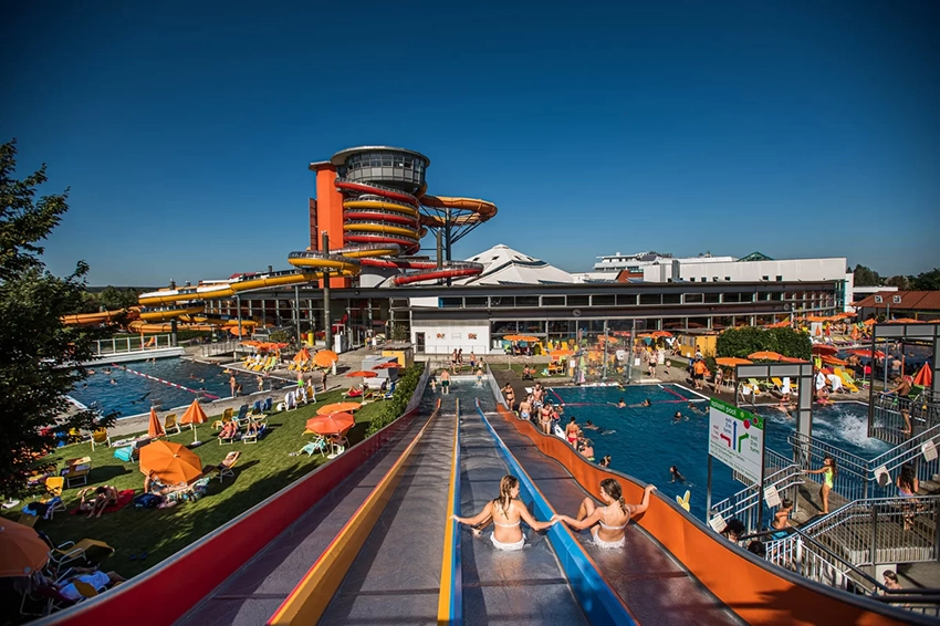 Sommertag in der Sonnentherme Lutzmannsburg mit Badegästen, die sich in mehreren Schwimmbecken entspannen und auf farbenfrohen Wasserrutschen Spaß haben