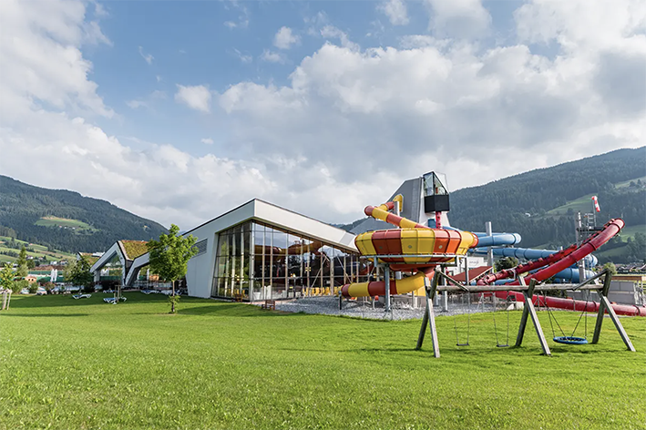 Außenansicht der Therme Amadé mit Röhrenrutschen, umgeben von einer grünen Wiese und malerischen Berglandschaften im Hintergrund, unter klarem Himmel